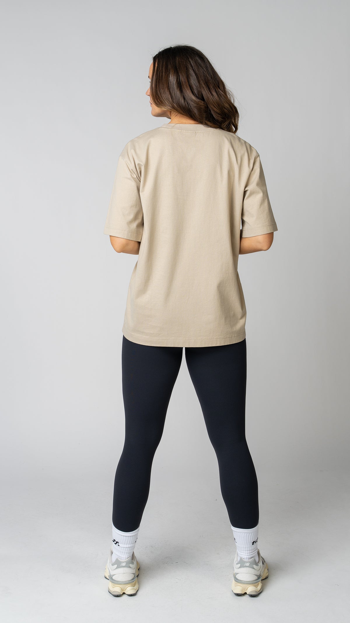 Woman wearing a beige t-shirt, black leggings, and white sneakers on a gray background. Back view.